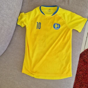 Sverige fotbollströja gul H&M nr 10 - Gul fotbollströja från H&M med blå detaljer och nummer 10 på både fram- och baksidan. Sverige-emblem på bröstet och texten 'Sverige' på ryggen. Tillverkad i 100% polyester, lätt och ventilerande material. Rund hals med blå kant och korta ärmar med blå kant.