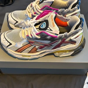 Balenciaga Runner sneakers multicolor - Säljer ett par Balenciaga Runner sneakers i storlek 44. Skorna har en futuristisk design med mesh och syntetmaterial i vitt, silver, grått, gult, lila och orange. De har chunky sula, snörning och coola detaljer som kontrastfärgade paneler och branding på både tunga och häl.