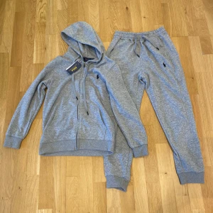 Polo Ralph Lauren zip hoodie och byxor - Säljer en grå ziphoodie från Polo Ralph Lauren med dragkedja och huva. Tröjan har snörning vid halsen, ribbade muddar och det klassiska Polo-logot broderat på bröstet. Även ett par lika byxor i samma färg.  Materialet är mjukt och skönt, perfekt för alla dagar. Custom fit-modell. Jag har aldrig använt de.