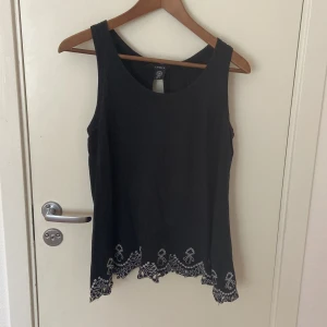 Svart broderad linnetopp från Lindex - Svart ärmlös linnetopp från Lindex med vit brodyr längs nederkanten och ryggen. Toppen har en snygg öppen detalj i ryggen och vågig kant nertill. Perfekt för dig som gillar unika detaljer och stilren design.
