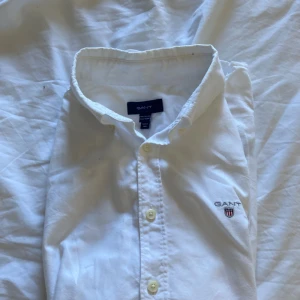Vit skjorta från Gant med broderad logga - Klassisk vit skjorta från Gant med broderad logga på bröstet. Skjortan har lång ärm, knappar framtill och en stilren krage. Tillverkad i bomull för en skön och luftig känsla. Perfekt för dig som gillar en clean och tidlös stil.