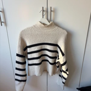 Randig stickad tröja från Bershka - Säljer en mysig stickad tröja från Bershka i storlek S. Tröjan är vit med svarta horisontella ränder, har en ribbad hög hals och långa ärmar. Croppad passform som ger en cool vibe till din outfit