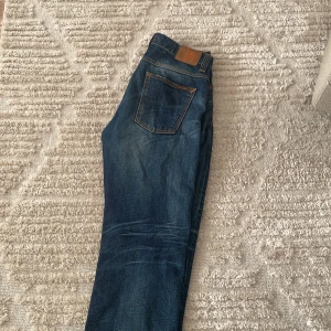 Nudie Jeans - Säljer ett par blå jeans från Nudie Jeans med snygga slitningar och hål på knäna. Klassisk femficksmodell med orangea sömmar och läderpatch bak i midjan. Jeansen har en rak passform och är tillverkade i robust denim. Säljs för 599 och har aldrig använt dem hör av er vid funderingar😀