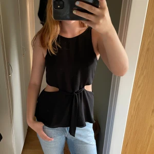 Svart ärmlös topp med knyt och cut-outs - Snygg svart ärmlös topp med cut-outs i midjan och knyt framtill. Toppen har rund halsringning och en cool v-formad rygg. Materialet är mjukt och faller snyggt, perfekt till jeans eller kjol för en trendig look. Gammal från zara