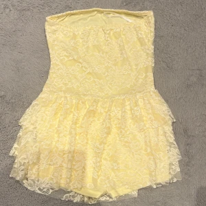 Ljusgul axelbandslös spetsklänning - Superfin ljusgul axelbandslös kortklänning med blommig spets över hela klänningen. Kjolen är volangig och ger en söt och romantisk vibe. Perfekt för sommarens alla tillfällen när du vill sticka ut lite extra.