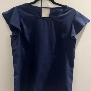Mörkblå blus med volangärm - En stilren mörkblå blus med rund halsringning och korta volangärmar. Toppen har en enkel, rak passform och en liten öppning i nacken. Perfekt för dig som gillar en clean look med en twist.