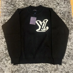 Svart stickad tröja Louis Vuitton - Svart stickad tröja från Louis Vuitton med stor vit LV-logga på bröstet. Tröjan har rund hals, ribbade muddar och lång ärm. Perfekt för dig som vill ha en clean och lyxig look med tydlig designer-vibe.