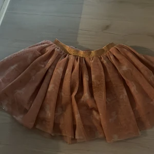 Glittrig rosa tyllkjol med guldresår - Supersöt rosa tyllkjol med glittrande detaljer och bredt guldigt resårband i midjan. Kjolen är fluffig och har flera lager för extra volym. Perfekt för dig som gillar att sticka ut med lite extra glitter och färg.