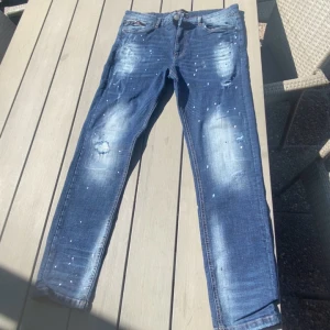 Blå jeans Supply & Demand XL - Snygga blå jeans från Supply & Demand med coola slitningar och ljusa tvättade partier. Jeansen har vita färgstänk och klassisk femficksdesign. Passformen är rak med normal midja och materialet är robust denim. Perfekt för dig som gillar streetstyle och vill sticka ut.