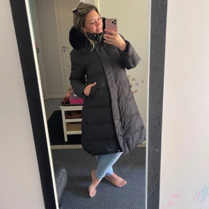 Svart lång pufferjacka med päls - Säljer en svart lång pufferjacka med fluffig svart päls runt huvan. Jackan har raka linjer, två sidofickor och stängs med dragkedja framtill. Perfekt för kalla dagar och har en stilren look som funkar till jeans och sneakers.Helt ny.Storlek motsvarar eu 40