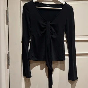 Svart omlottopp med knyt från Gina Tricot - Svart långärmad omlottopp från Gina Tricot i storlek XS. Toppen har v-ringning och snygg knytning framtill som ger en cool detalj. Materialet är mjukt och stretchigt, perfekt för att skapa en trendig look. Passformen är slim och toppen är croppad. Original pris 300kr.