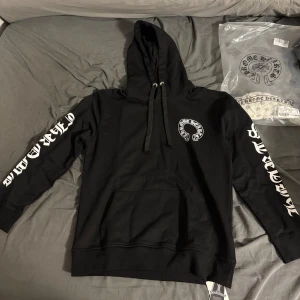 Chrome Hearts svart hoodie med tryck - Svart hoodie från Chrome Hearts med stor vit logga på ryggen och mindre logga på bröstet. Snygga vita tryck längs båda ärmarna och coolt rött och vitt läppmotiv på ena ärmen. Klassisk huva med snörning och stor magficka framtill. Perfekt för dig som gillar streetwear.