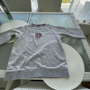 Grå sweatshirt från GANT med logga - Säljer en grå sweatshirt från GANT med broderad logga och emblem på bröstet. Tröjan har lång ärm, rund hals och ribbade muddar. Materialet är mjuk bomull, perfekt för chill dagar. Passar dig som gillar klassisk och sportig stil.