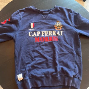 Blå sweatshirt från Morris Cap Ferrat - Snygg marinblå sweatshirt från.med broderade detaljer och text i vitt och rött på bröstet. Tröjan har rund hals, långa ärmar och ribbade muddar.