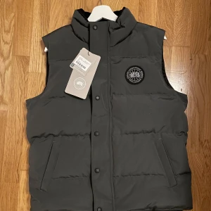 Svart dunväst från Canada Goose - Säljer en svart dunväst från Canada Goose med hög krage och snyggt broderat logomärke på bröstet. Västen har raka linjer, två sidofickor och knäppning med svarta knappar framtill. Perfekt för kalla dagar när du vill hålla stilen.