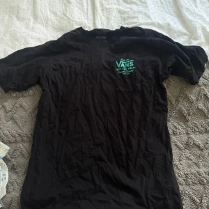 Svart Vans t-shirt med turkos tryck - Svart t-shirt från Vans i klassisk passform, tillverkad i 100% bomull. Framtill och baktill finns ett stort turkost Vans-tryck med texten 'Off The Wall Custom Classics'. Rund hals och korta ärmar, perfekt för dig som gillar streetwear.