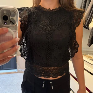 Svart spetsblus med volangdetaljer - Svart blus i spets med volangdetaljer längs ärmar och nederkant. Knappt använd. 