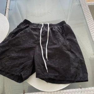 Svarta Nike shorts med snörning M - Svarta shorts från Nike i storlek M med vit snörning i midjan och diskret mönster med Nike-loggor över hela tyget. Shortsen har elastisk midja och innerbyxa i mesh för extra komfort. Perfekta för sport eller chill dagar.