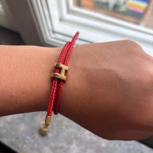 Rött Hermès armband med H-detalj - Snyggt rött armband från Hermès med dubbla flätade band och en ikonisk H-detalj i guldfärgad metall. Avslutas med guldfärgade ändar för en lyxig touch. Perfekt accessoar för att lyfta din stil och ge en färgklick till handleden.