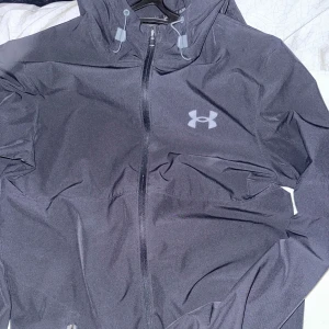 Svart vindjacka från Under Armour - Svart vindjacka från Under Armour med stilren design och logga på bröstet. Jackan har hel dragkedja framtill, två sidofickor med dragkedja och långärmade ärmar. Perfekt för aktiva dagar och sportiga outfits.