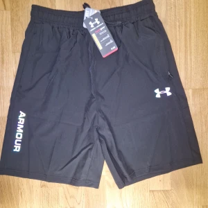 Svarta träningsshorts Under Armour - M men kan passa L. Snygga svarta träningsshorts från Under Armour med logga på benet och elastisk midja. Perfekta för gymmet eller löprundan, med lätt och bekvämt material som andas. Klassisk passform och diskret design gör dem enkla att matcha med din träningsoutfit.