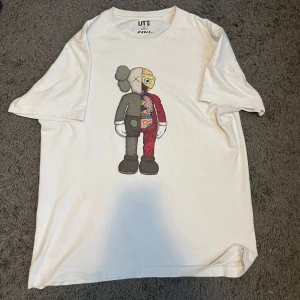 Vit KAWS x Uniqlo t-shirt - Vit t-shirt från Uniqlo UT i samarbete med KAWS. Framsidan har en stor tryckt KAWS Companion-figur i färg. Klassisk rund hals och korta ärmar. Materialet är mjuk bomull, perfekt för dig som gillar streetwear och konstnärliga prints.
