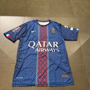 PSG matchtröja D.Doué #14 Nike blå - Paris Saint-Germain fotbollströja med D.Doué och nummer 14 på ryggen. Tröjan är marinblå med röda och vita detaljer, har ett geometriskt mönster framtill och Champions League-patch på ärmen. Tillverkad i lätt polyester med Dri-FIT-teknologi för bästa komfort.