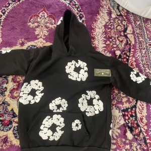 Svart hoodie från Denim Tears med tryck - Svart hoodie från Denim Tears med stora vita grafiska tryck i form av cirklar över hela tröjan. Tröjan har huva, känguruficka och en patch med logga på bröstet. Materialet känns som mjuk bomull och passformen är normal. Perfekt för dig som gillar streetwear och unika prints.