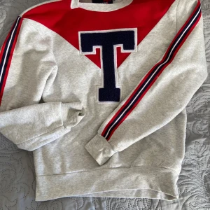 Grå sweatshirt med röd detalj och T -  grå sweatshirt med röd trekant upptill och ett stort mörkblått T på bröstet. Ärmarna har rödblå ränder längs sidorna. Mjuk och ribbade muddar vid ärmslut och nederkant.