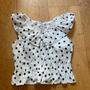 Prickig volangblus från Zara - Supersöt vit blus från Zara med svarta prickar och bred volangkrage. Ärmlös modell med smock i midjan och volang nertill som ger en riktigt gullig och trendig look. Perfekt för dig som gillar lekfulla detaljer och vill sticka ut lite extra.
