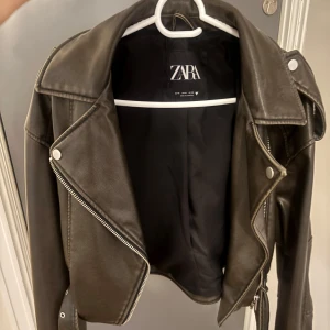 Oanvänd populär skinnjacka från Zara - Snygg svart skinnjacka från Zara med klassisk bikerstil. Jackan har silvriga dragkedjor, breda slag och bälte i midjan. Croppad modell som ger en cool vibe och passar perfekt till jeans eller klänning. Fodrad insida och detaljerade sömmar för extra edge. 