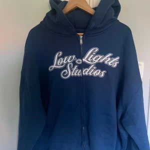 Blå hoodie från Low Lights Studios - Snygg blå hoodie från Low Lights Studios med broderad vit text på bröstet. Tröjan har huva, dragkedja framtill och är tillverkad i mjuk bomull. Perfekt för chill dagar och streetwear.          Helt nyskick.                                                              Pris kan diskuteras!                                                    Nypris runt 1100kr.                        