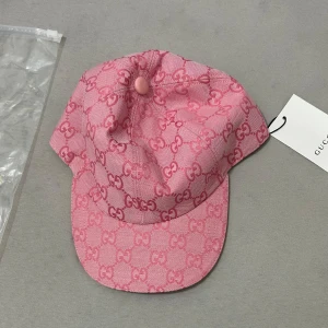 Rosa Gucci keps  - Snygg rosa keps från Gucci med ikoniskt GG-mönster över hela kepsen. Justerbar rem baktill i rosa och detaljerad med en liten metallknapp på toppen. Materialet är bomull och kepsen har en klassisk böjd skärm. Perfekt för dig som vill sticka ut med lyxig streetstyle.