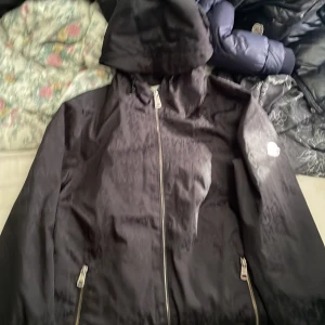 Svart Moncler vindjacka med huva - Svart vindjacka från Moncler med huva och hel dragkedja framtill. Jackan har diskret Moncler-logga i mönstret över hela tyget och ett broderat Moncler-märke på ärmen. Två fickor med dragkedja och coolt serietryck på insidan. Perfekt för dig som gillar street och premium. inte trasig bra skick vet inte om den är äkta köpt av en vän för 3500 aldrig andvänd för den e för stor bara testat 