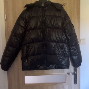 Svart Moncler pufferjacka med huva - Svart pufferjacka från Moncler med glansig finish och huva. Jackan har dragkedja framtill, två sidofickor med dragkedja och klassisk Moncler-logga på ärmen. Perfekt för kalla dagar och har en riktigt schysst oversized look.