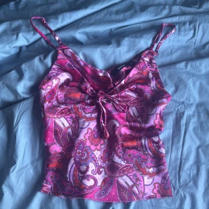 Paisleymönstrad rosa linnetopp BikBok - Säljer en färgstark linnetopp från BikBok i storlek XS. Toppen har ett paisleyinspirerat mönster i olika nyanser av rosa, lila och rött, tunna axelband och en snygg knytning framtill. 