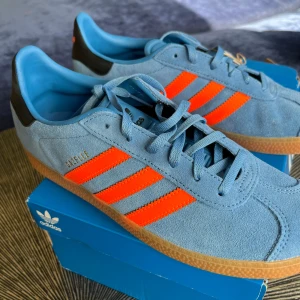 Adidas Gazelle blå och orange sneakers - Säljer dessa snygga ljusblå Adidas Gazelle sneakers med orange ränder. Skorna är aldrig använda och har prislappen kvar💙