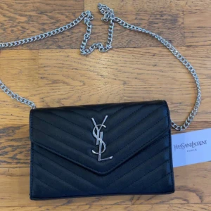 Ysl väska  - Snygg svart axelväska från Yves Saint Laurent med silverfärgad kedja och ikonisk YSL-logga framtill. Väskan har quiltad design och är tillverkad i skinn. Perfekt storlek för det viktigaste och ger en lyxig vibe till din outfit.