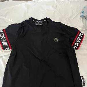 Svart Philipp Plein t-shirt med logga - Svart t-shirt från Philipp Plein med coola logoband i vitt och rött på ärmarna. Klassisk rund hals och en diskret PP-logga på bröstet. Materialet är mjuk bomull och passformen är normal. Perfekt för dig som gillar streetwear med lyxig vibe.