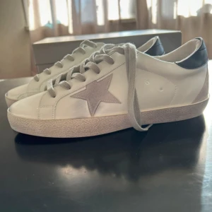 Golden Goose Superstar sneakers vit/grå - Golden Goose Superstar sneakers i vitt skinn med grå mockadetaljer och en ikonisk stjärna på sidan. Skorna har grå snörning, svart hälparti med logga och platt sula. Perfekt för dig som gillar stilrena och trendiga sneakers med unika detaljer.