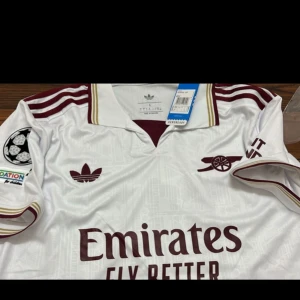 Arsenal borta fotbollströja med Gyökeres 14 - Snygg Arsenal fotbollströja i vit med vinröda detaljer och tryck. Tröjan har klassisk krage, korta ärmar och Champions League-märke på ena ärmen. Gyökeres och nummer 14 på ryggen. Helt ny och oanvänd 