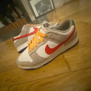 Nike Dunk Low med röd swoosh - Nike Dunk Low sneakers med vit och beige ovandel i skinn och mocka, röd swoosh på sidan och orange snören. Små gula och gröna detaljer på hälen och tå. Perfekt för dig som gillar färg och streetstyle storlek 40,5.