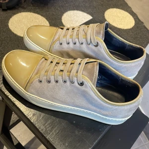 Lanvin sneakers i beige mocka - Säljer ett par snygga Lanvin sneakers i beige mocka med glansiga ljusbruna tåpartier och matchande snörning. Skorna har rund tå, platt vit sula och detaljer i skinn runt öppningen. Perfekt för dig som gillar stilrena och lyxiga sneakers.priset kan sänkas vid snabb affär 
