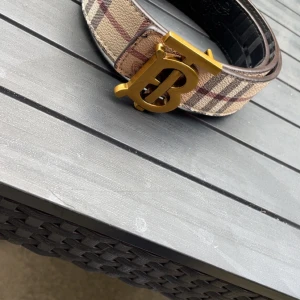 Burberry rutig bälte med guldspänne - Snyggt Burberry bälte med klassiskt rutigt mönster i beige, brun, röd och grå. Bältet har ett stort guldigt metallspänne med Burberrys ikoniska TB-logga. Materialet är skinn med textil på ovansidan och detaljerade Burberry-gravyrer. Perfekt för att lyfta din outfit. 115 lång