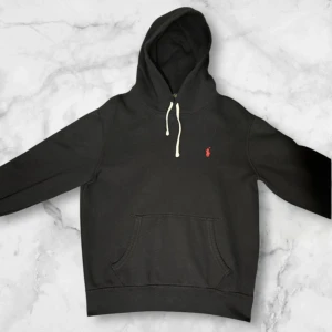 Svart hoodie från Polo Ralph Lauren - Svart hoodie från Polo Ralph Lauren med klassisk röd logobrodering på bröstet. Tröjan har huva med vita snören, stor magficka och ribbade muddar. Mjuk insida i bomullsmix, perfekt för chill dagar. Passformen är normal och ärmarna är långa. Bra skick inga defekter.