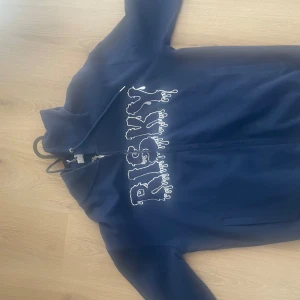 Blå hoodiejacka från Risky, S - Snygg mörkblå hoodiejacka från Risky med stort broderat logotyptryck i vitt på bröstet. Jackan har dragkedja framtill, huva med snörning och mjukt bomullsmaterial. Perfekt för dig som gillar streetwear och vill sticka ut med coola detaljer.