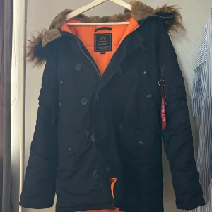 Svart jacka från Alpha Industries M - Jacka från Alpha Industries i svart med orange foder och stor huva med brun fuskpäls. Jackan har både dragkedja och knappar, samt en ikonisk röd 'Remove Before Flight'-tag på ärmen. Flera fickor och detaljer som ger en cool streetkänsla. Perfekt för kalla dagar.