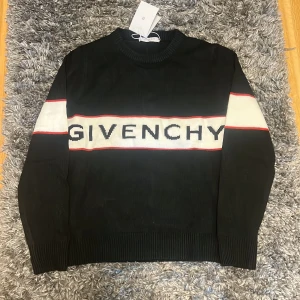 Svart stickad tröja från Givenchy - Svart stickad tröja från Givenchy med vit och röd rand över bröstet och ärmarna. Stor Givenchy-logga framtill. Rund hals och ribbade muddar vid ärmslut och nederkant. Perfekt för dig som gillar exklusiva märken och stilren design.