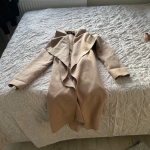 Beige kappa från Esmara med bälte - Snygg beige kappa från Esmara med långa ärmar och ett brett, vattenfallsinspirerat slag. Kappan har ett matchande bälte i midjan för en clean och stilren look. Perfekt för dig som vill ha en klassisk och trendig vibe till höst och vår.