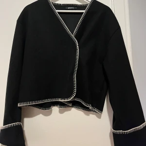 Svart omlottkofta från Gina Tricot - Svart omlottkofta/tunnare jacka från Gina Tricot med vita dekorativa sömmar längs kanterna. Knappast använd, pris kan diskuteras🤎